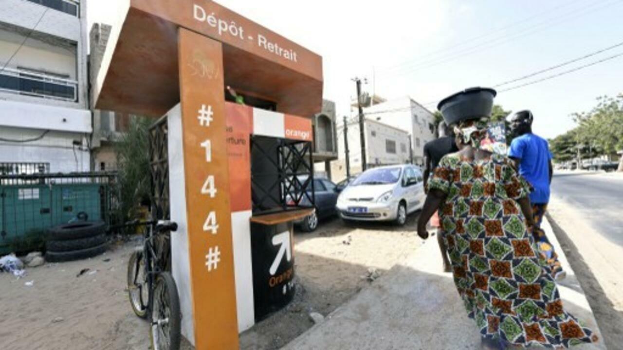 Customers pass in front of an « Orange Money » booth, on July 7, 2015 in Ouakam, a suburb of Dakar. These booths run by the Sonatel Group and owned 42% by France Telecom, are used to transfer money by telephone and are installed around Senegal. They have created jobs for the youth and have been received enthusiastically by users. AFP PHOTO / SEYLLOU / AFP PHOTO / SEYLLOU